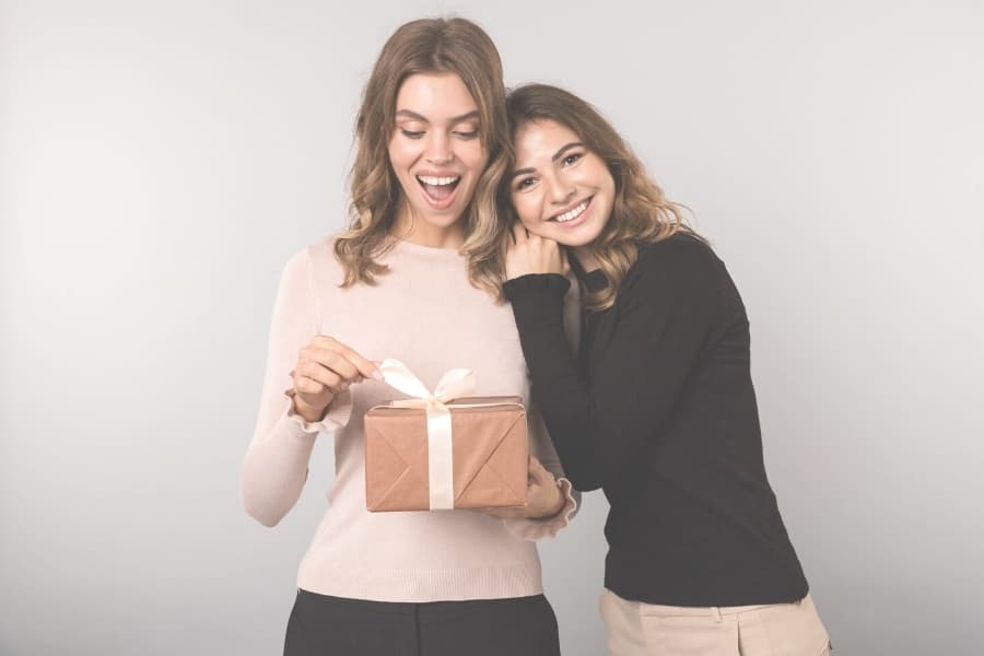 Two women posing. One woman in a pink shirt is opening up a present.